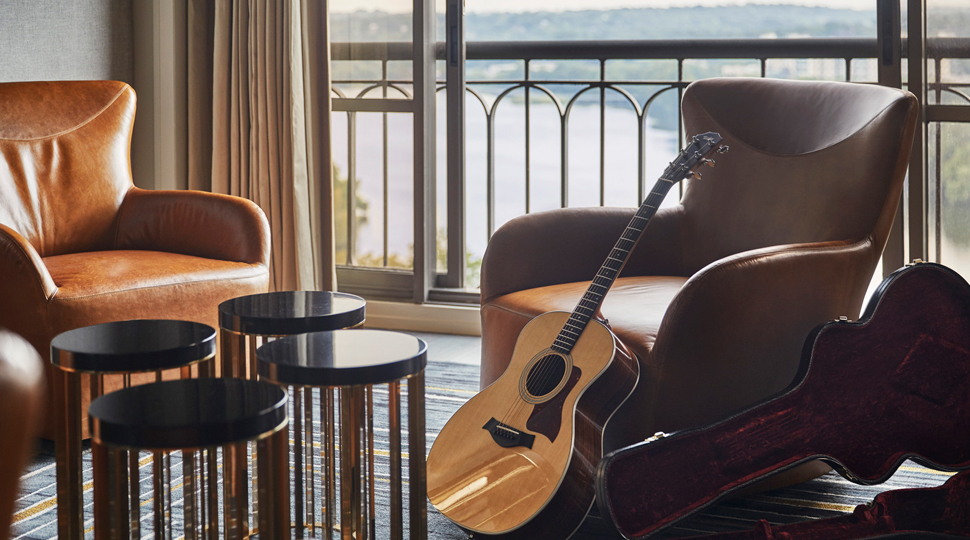 An acoustic guitar leans against a brown leather arm chair with four small round, black tables and another brown leather arm chair on the other side of the room and a balcony in the background
