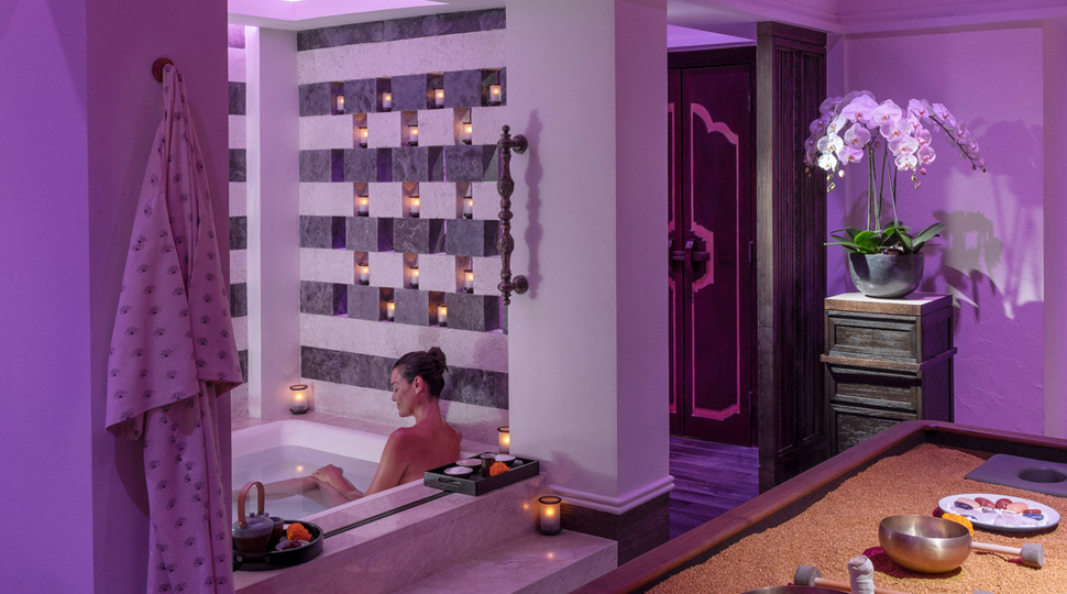 Person lounges in a bathtub set within a private spa suite and lit with purple light