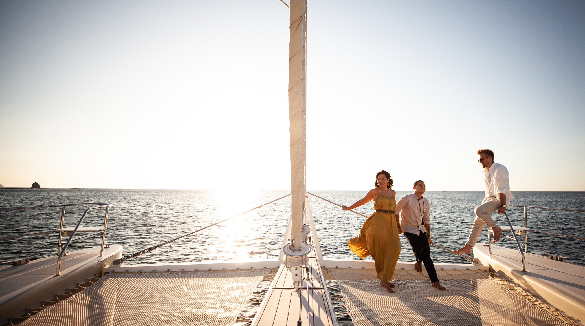 Three people stand on the edge of a sail boat as the sun sets behind them