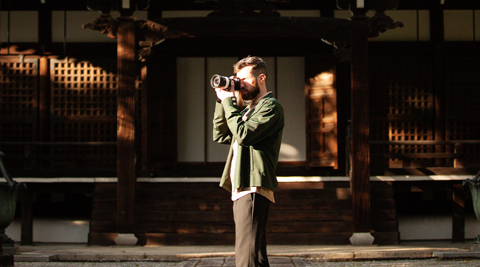 Person wearing olive green jacket and brown pants stands looking through a camera