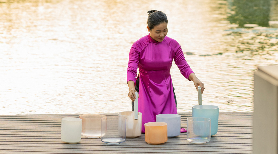 A person wearing a bright magenta robe kneels in front of eight crystal bowls set out on a wooden deck by the water