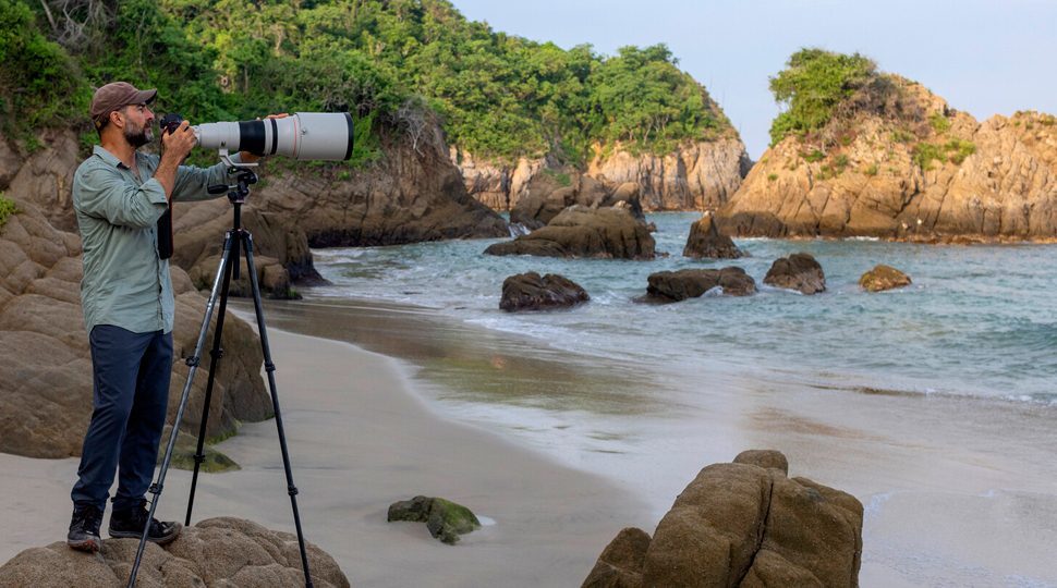 Photographer stands behind a large macro lens on a rocky beach